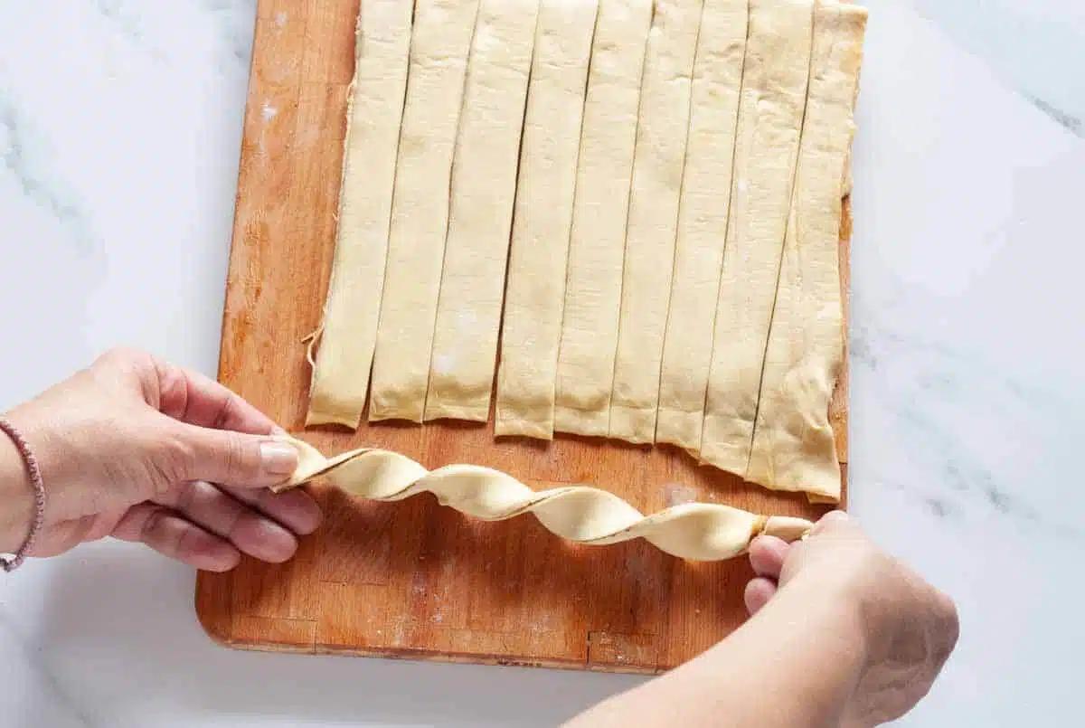 hands twisting strips of puff pastry dough on a wooden board