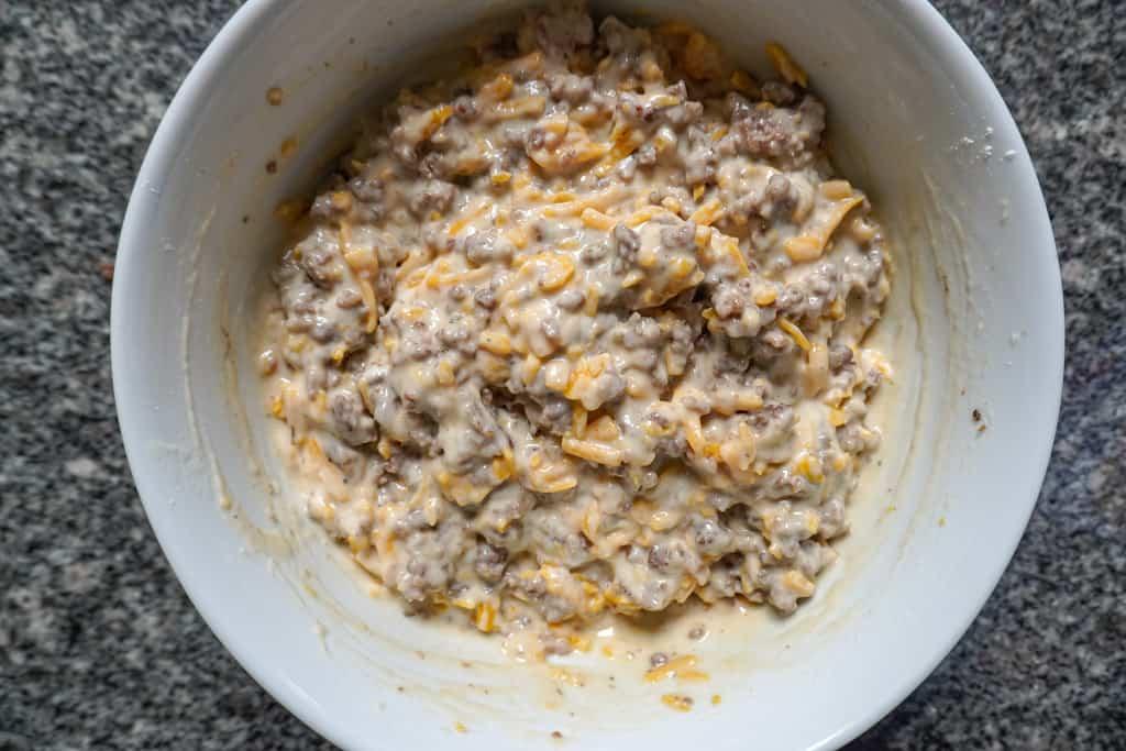 Hands gently placing small, cooked breakfast sausage crumbles into pancake batter in a mixing bowl