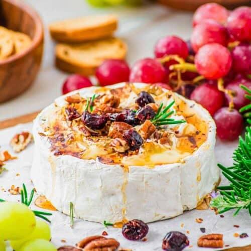 Gourmet baked brie wheel oozing with honey and surrounded by crackers and fresh fruit on a festive platter, cozy winter setting