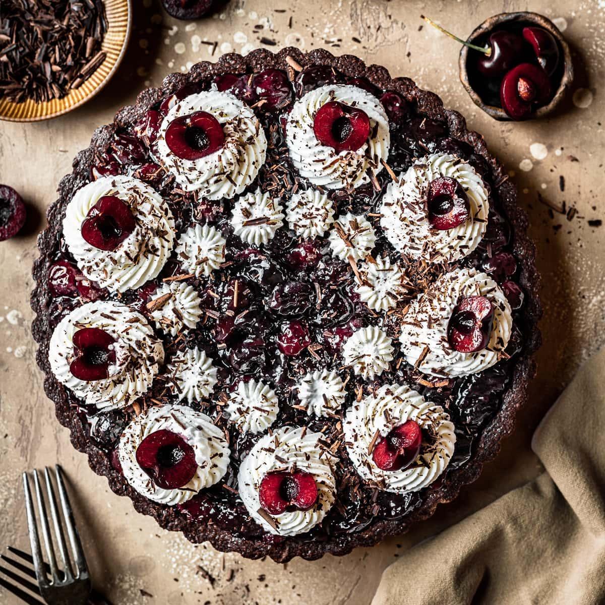 A whole cherry chocolate tart on a rustic wooden table with some fresh cherries scattered around it