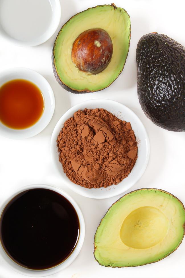 Overhead shot of the ingredients for avocado chocolate mousse arranged on a wooden surface, including ripe avocados, cocoa powder, chocolate, and maple syrup.