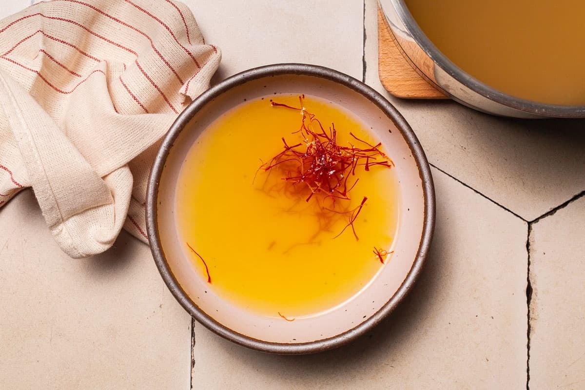Overhead shot of saffron threads being steeped in warm broth, with a wooden spoon nearby