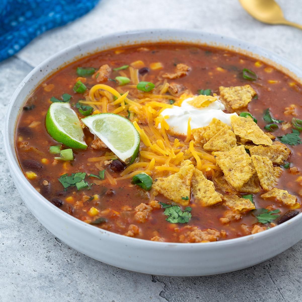 A steaming bowl of creamy taco soup with a dollop of sour cream, shredded cheese, and fresh cilantro on top, with tortilla chips on the side, cozy kitchen background