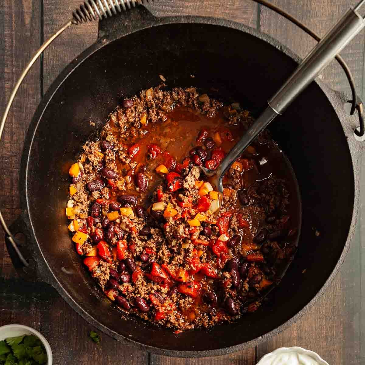 Dutch oven cooking bold campfire chili over an open fire, rich steam rising