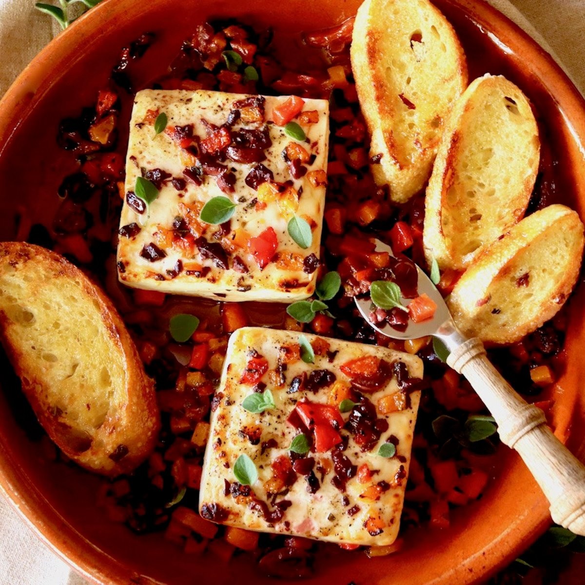 Close-up of baked feta cheese block in a baking dish, drizzled with olive oil and garnished with fresh oregano and red pepper flakes, served with crusty bread.