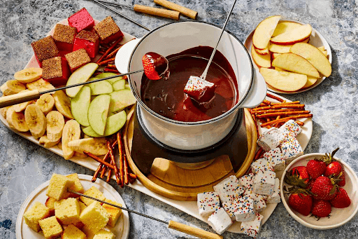 A decadent chocolate fondue with various fruits and treats arranged around the fondue pot.