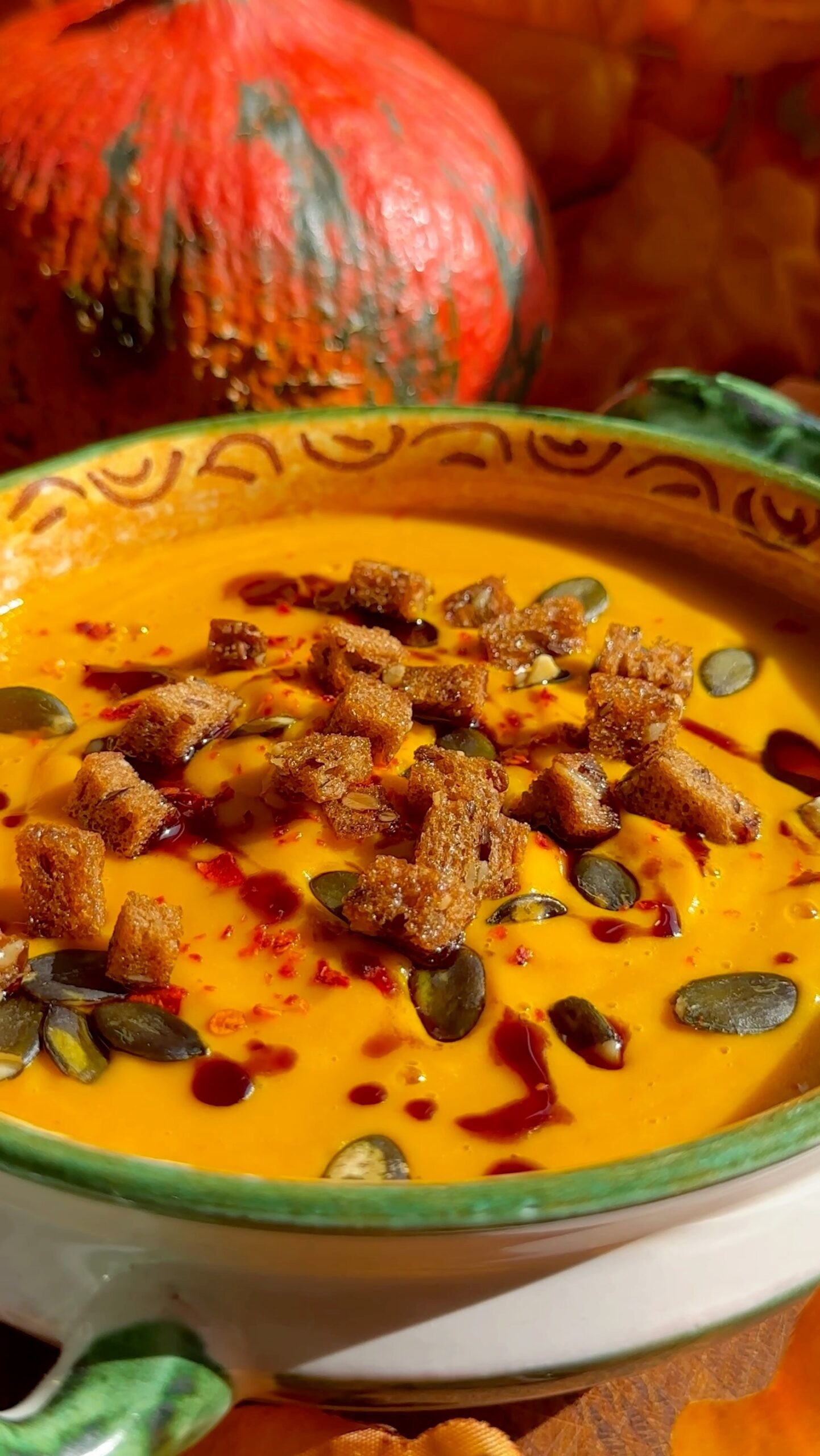 A rustic bowl of spiced pumpkin soup garnished with toasted pepitas, fall leaves in the background