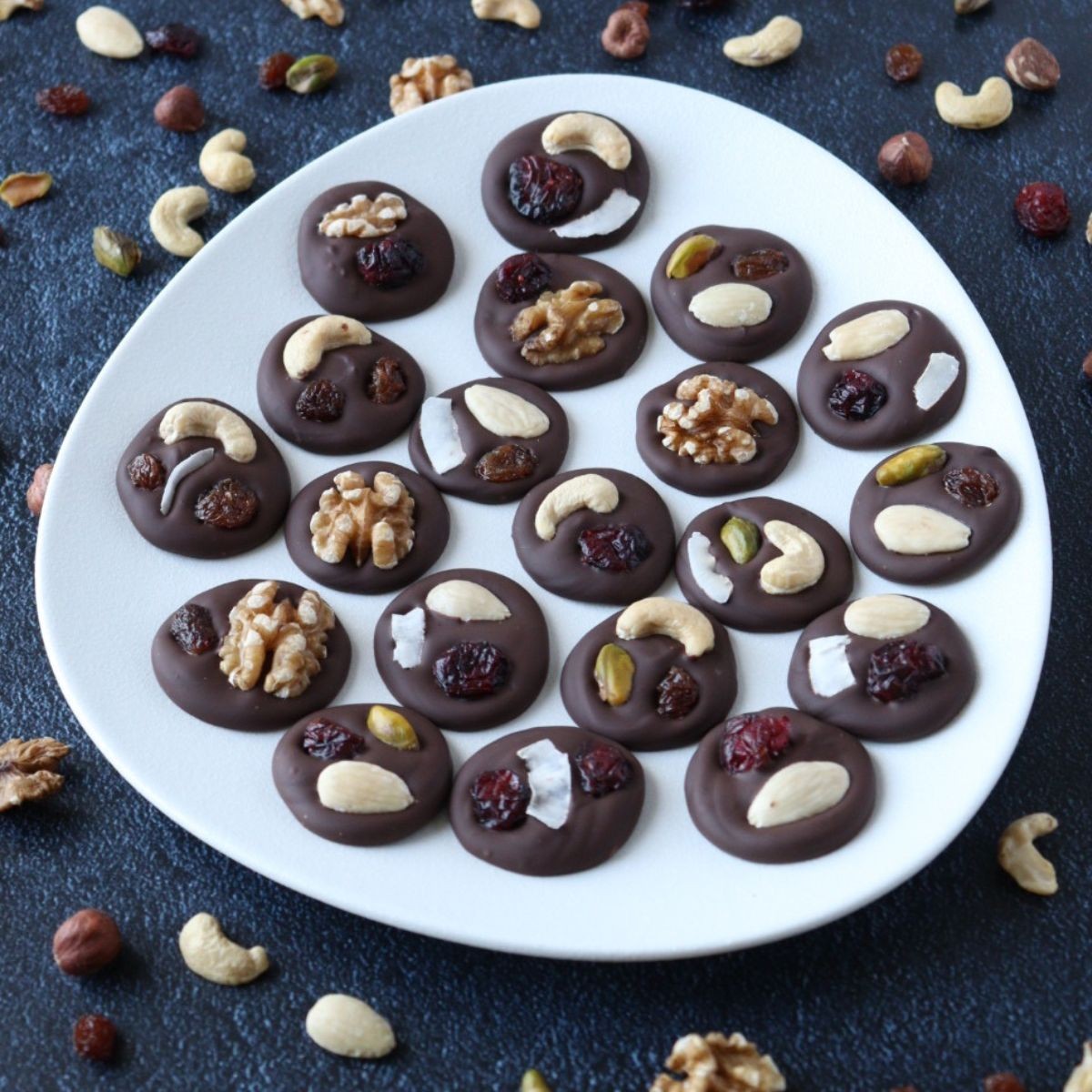 elegant dark chocolate mendiants with various dried fruits and nuts on a white platter, gourmet look