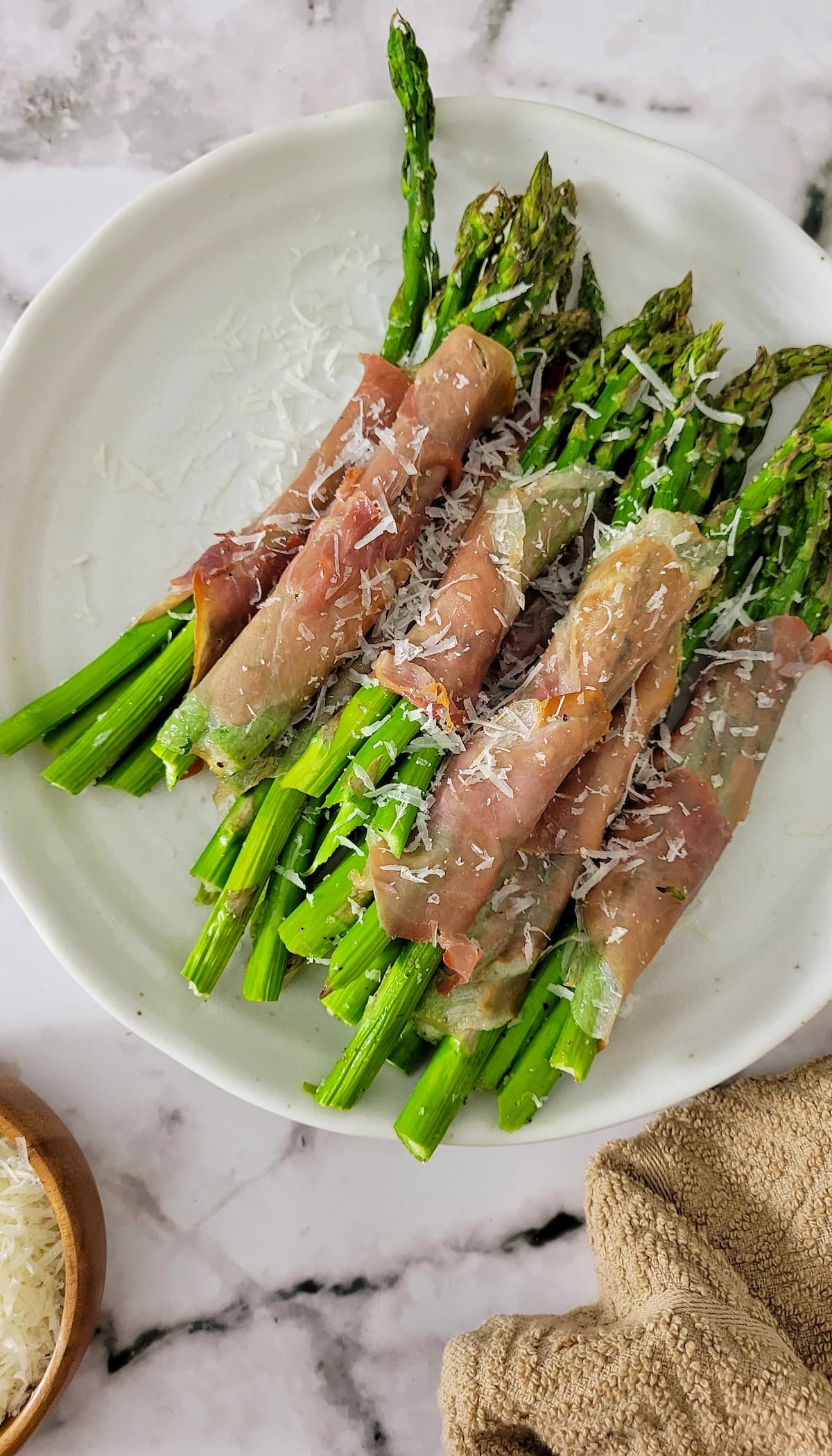Crispy prosciutto wrapped asparagus bundles on a white platter, garnished with fresh herbs