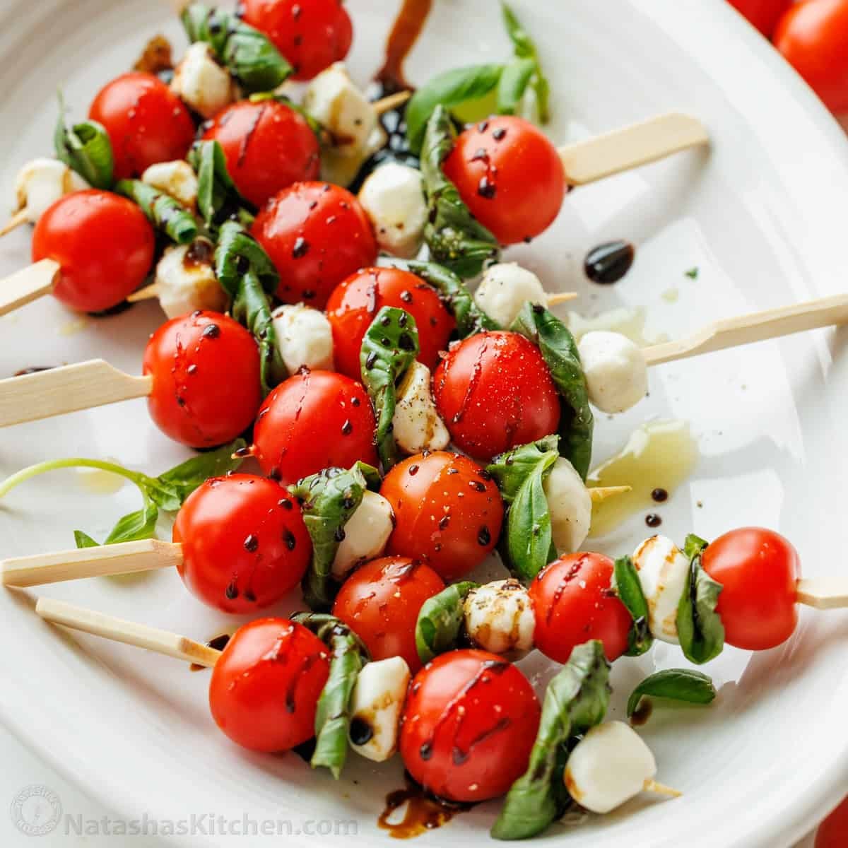 vibrant caprese skewers on a white platter with a drizzle of balsamic glaze, basil leaves, and grape tomatoes in soft natural light