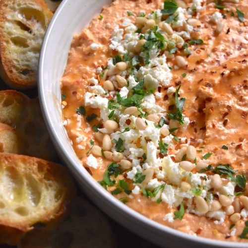 A vibrant bowl of roasted red pepper dip with feta cheese and olives, garnished with fresh herbs
