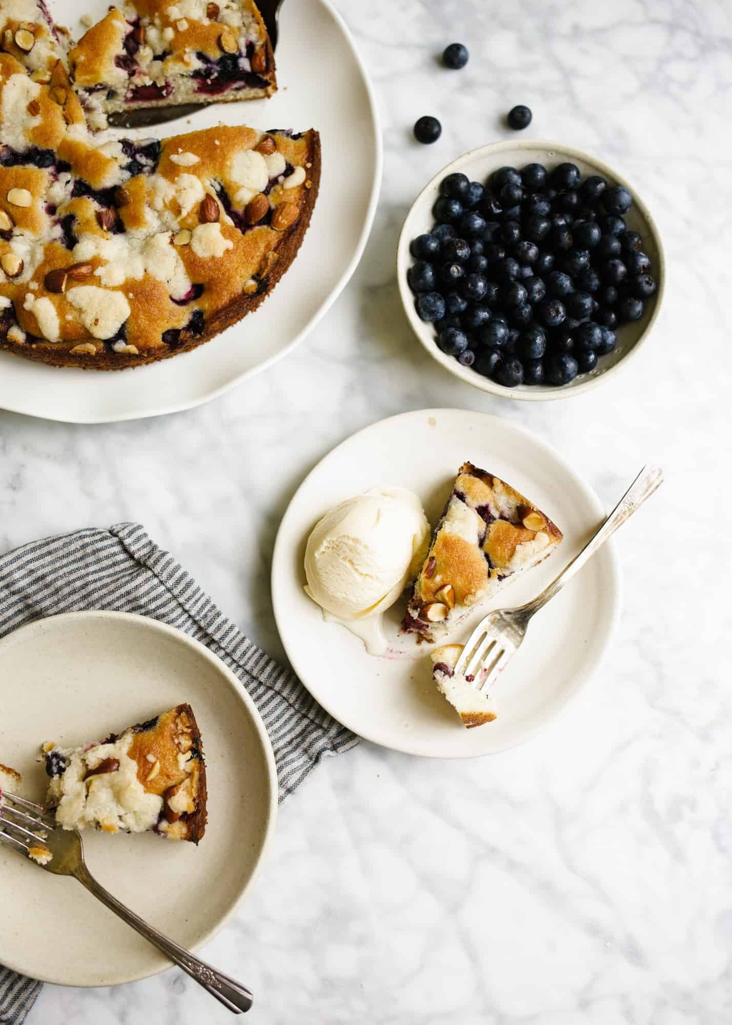 A rustic wooden table with a golden-brown wild berry buckle, with berries bubbling on top, steam rising, and a scoop of vanilla ice cream melting on top